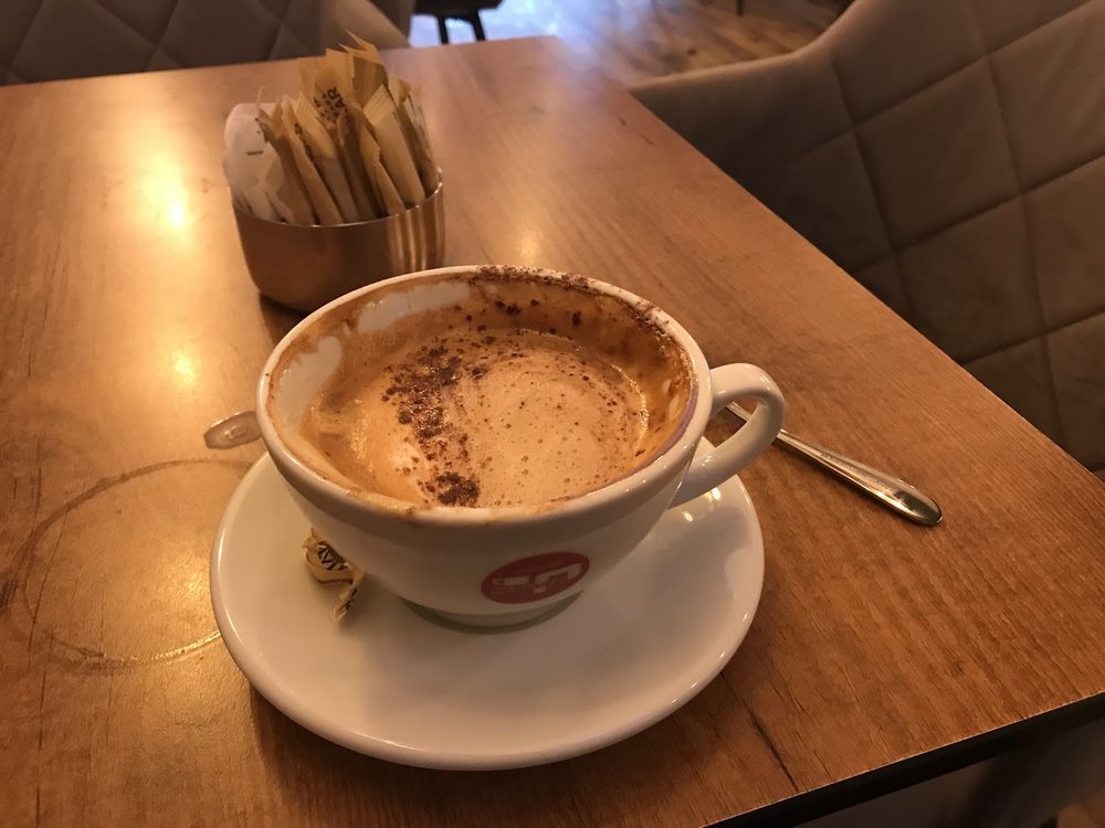 A chocolate-dusted cappuccino on a saucer beside a caddy of sugar sticks, in a padded booth